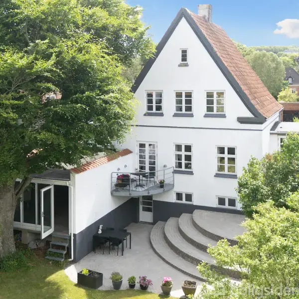 Hvidt, to-etagers hus med skråt tag står i en frodig have. Rund terrasse foran huset med stole og bord. Stort træ til venstre og blå himmel i baggrunden.