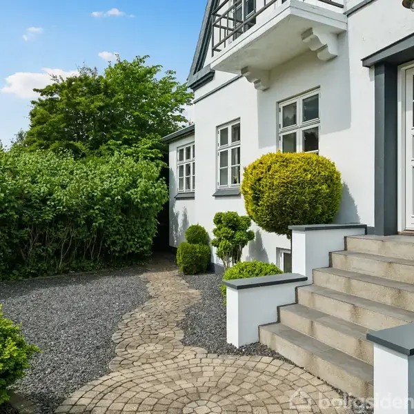 Husentré med en hvid dør og trapper, omgivet af trimmede buske. Stenbelagt sti fører gennem en anlagt have med grus og grønne træer i baggrunden.