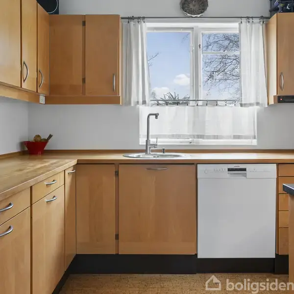 Køkken i træfinish har skabe og skuffer, omgivet af en opvaskemaskine og håndvask. Et vindue med udsigt lukker naturligt lys ind. Krydderier står på bordpladen i en rød skål.