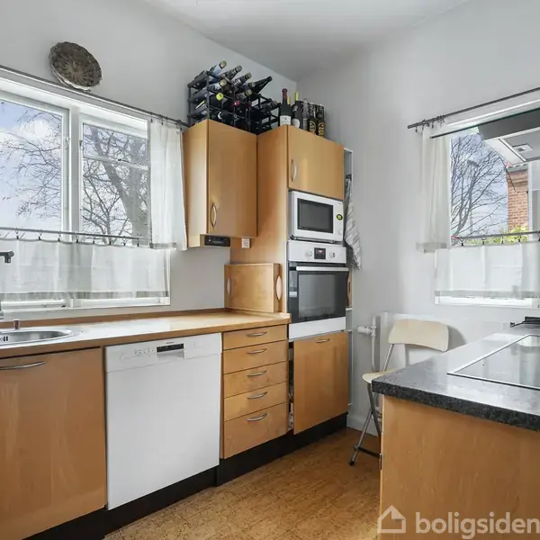 Et køkken med træskabe, en indbygget ovn og mikroovn. Der er en stor håndvask under et vindue, og kaffemaskine ved kogepladen. Vinduerne har korte gardiner, og der er trægulv.