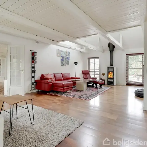 Rød sofa og lænestol står på tæppe nær en brændeovn i en lys stue med trægulv, hvide vægge og loftsbjælker. Trappe fører til overetagen.