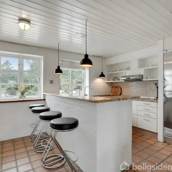 En lys køkken med en bardisk og fire moderne barstole på højre side. Køkkenet har hvidmalede vægge, hængende lamper, og en stor vindue, der giver naturligt lys.