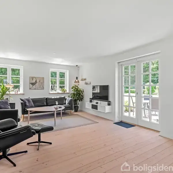 Moderne stue med sort læderstol og sofa, sofabord, TV ved væggen og store vinduer, der giver udsigt til en terrasse. Lys og enkel indretning med trægulv og planter.
