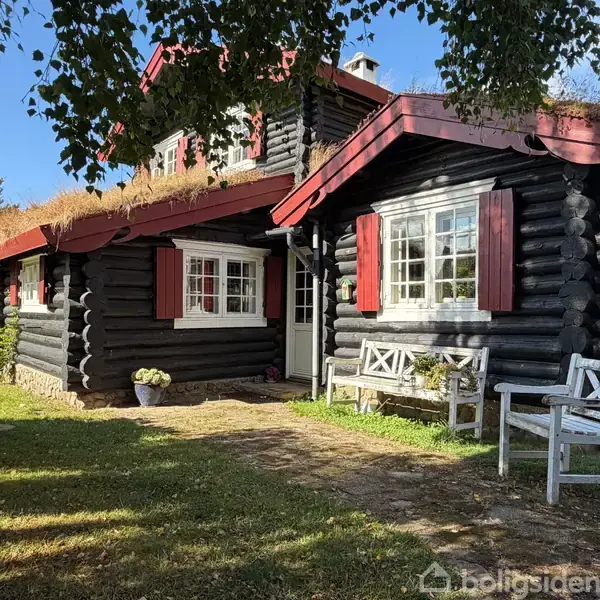 Et sort bjælkehus med røde skodder og græstag står i en grøn have. En hvid bænk og en stol er placeret foran huset, omgivet af træer.
