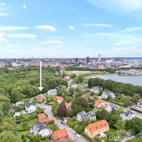 En hvid pil peger på et hus blandt flere huse med røde tage i en frodig forstad. I baggrunden ses en stor by med højhuse og en havn.