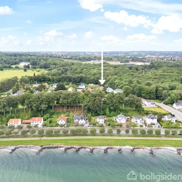 En række huse står langs en vej ved en strandpromenade. En pil peger mod et hus blandt træerne mod baggrunden. Området er naturskønt med både strand og skov.