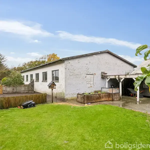 Et hvidt hus med fladt tag står stille i en grøn have med græs. En træterrasse er ved siden af huset, og der er buske og træer i baggrunden.
