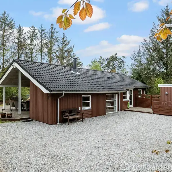 Et brunt træsommerhus med fladt tag står i en skovklædt have. Der er en grusbelagt indkørsel foran bygningen. Husets terrasser og træer omgiver huset.