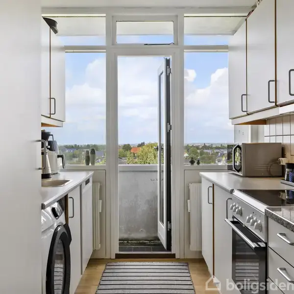 Et smalt køkken har hvide skabe og apparater, med en udsigt gennem en glasdør til en balkon og trætoppe udenfor. Teppets striber på gulvet tilføjer kontrast.