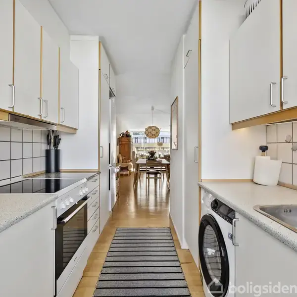 Et smalt, hvidt køkken med langsstillede skabe og apparater. En vask og vaskemaskine til højre, en komfur til venstre. En løber fører til en spisestue i baggrunden.