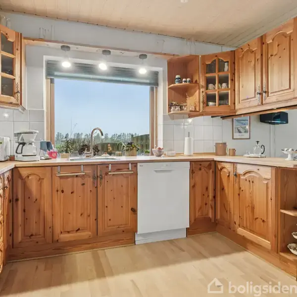 Køkken med træskabe arrangerer hylder og opvaskemaskine, lys strømmer ind gennem stort vindue med grøn udsigt, køkkenmaskiner står på bordplade; lys trægulv fuldender rummelig indretning.