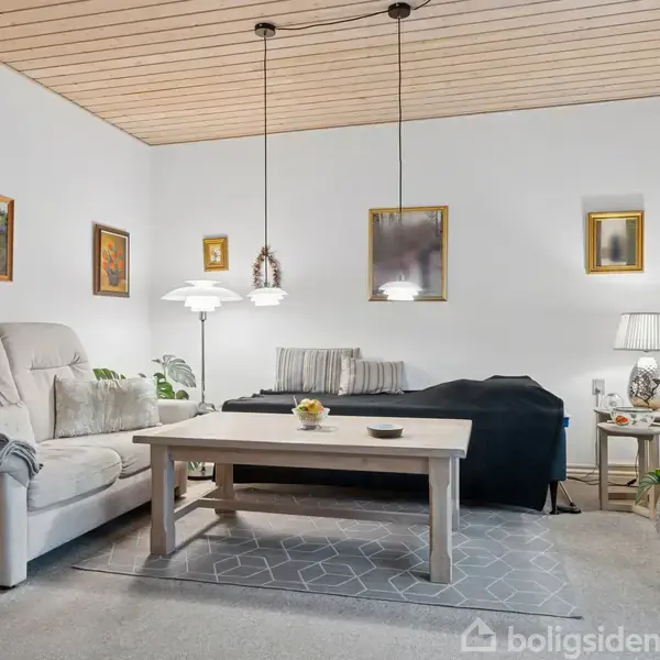 En lys stue med en beige sofa ved siden af et sofabord. En sort dækket sofa står bag bordet. Loftslamper hænger over bordet. Vægge prydes af kunstværker. Grønne planter i hjørnerne.