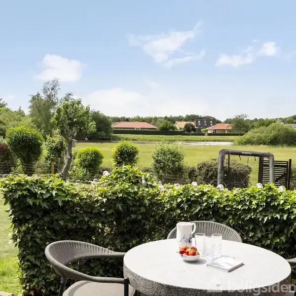 Rundt havebord med to stole står på terrasse. På bordet er en kande og skål med frugt. Bagved ses en grøn have omgivet af hække og en udsigt til marker.