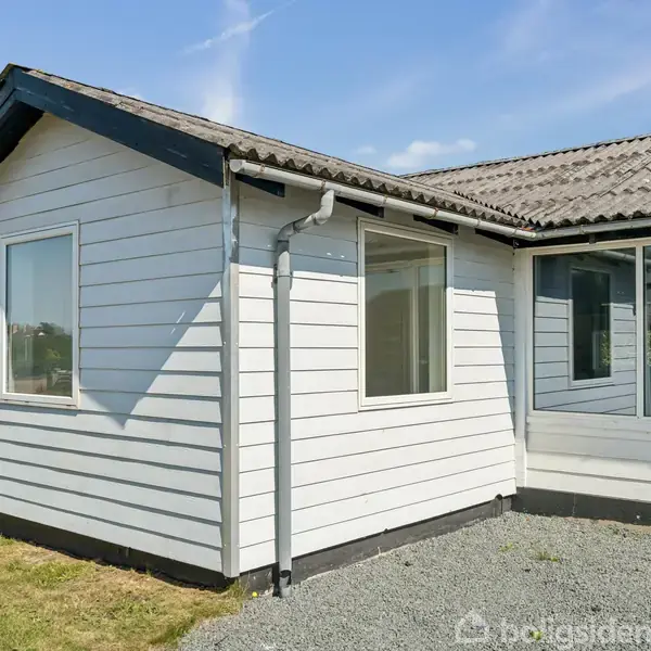 Et hvidt træhus med store vinduer står i et åbent område. Taget er skråt og beklædt med mørke fliser. Omkring huset er der en grusbelagt indkørsel og en græsplæne.