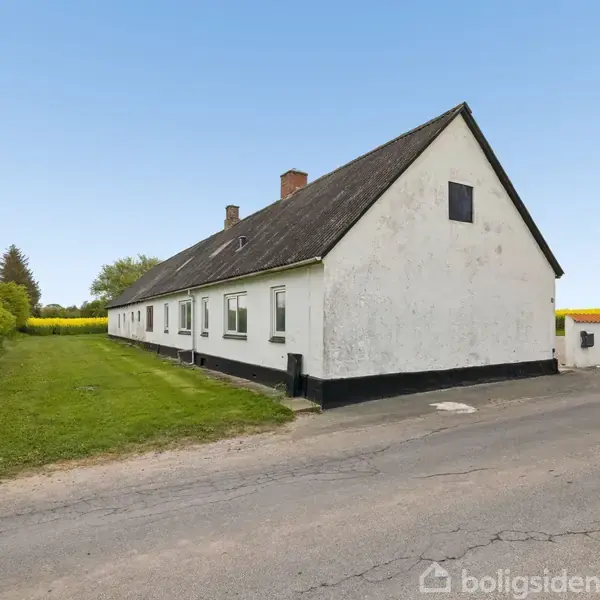 En lang hvid bygning med sort sokkel står ved en landevej. Græsplæne og træer omkranser huset på den ene side under en blå himmel.