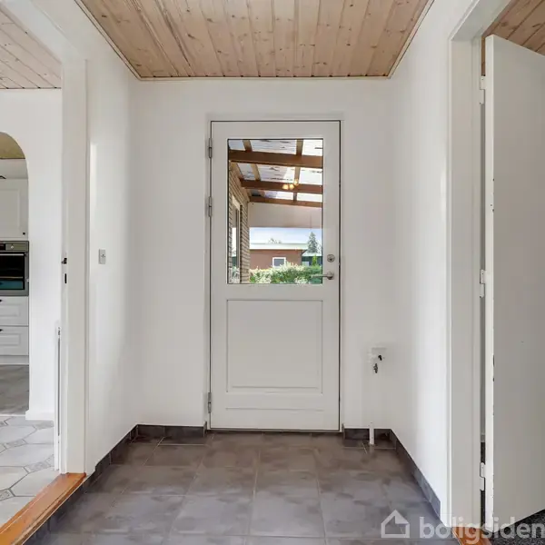 En hvid entré med flisegulv fører til en glasdør, der viser ud til en overdækket terrasse med udsigt til grønne træer. Værelser med åbne døråbninger ses til venstre og højre.