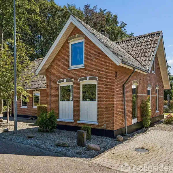 Et rødt murstenshus med hvidrammede vinduer står i et solbeskinnet, grønt område med træer og buske. Belægning af grus og brosten ses omkring huset.