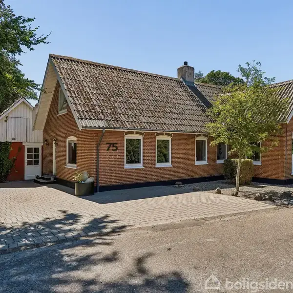En murstensvilla med gråt tag har nummeret "75" på facaden. Den står på en flisebelagt indkørsel, omgivet af træer, og har en tilstødende bygning til venstre.