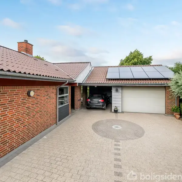 Rød murstensvilla med solpaneler på taget. En bil holder i indkørslen nær en garage med hvid port. Indkørslen er flisebelagt, omgivet af grønne hække og træer. Nummer 2 ses ved døren.
