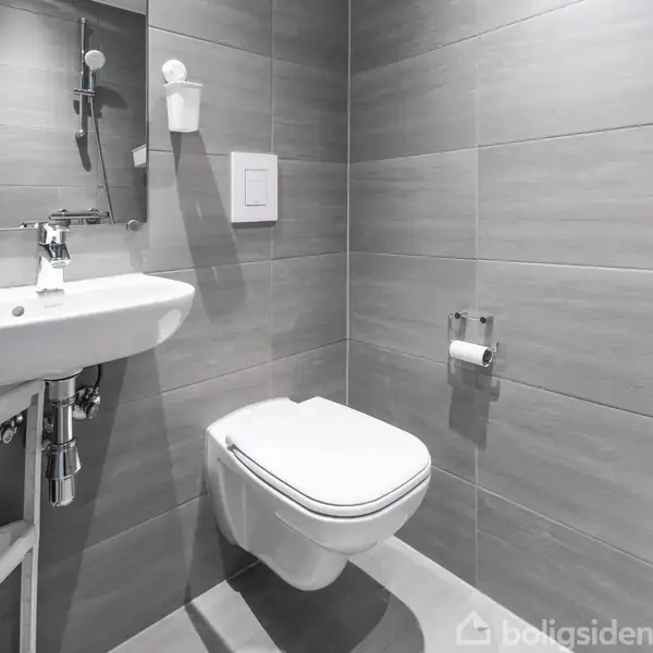 Toilet hænger på væggen i et moderne badeværelse med grå fliser. Ved siden af er en håndvask med et spejl over. Toiletpapir er monteret på væggen.