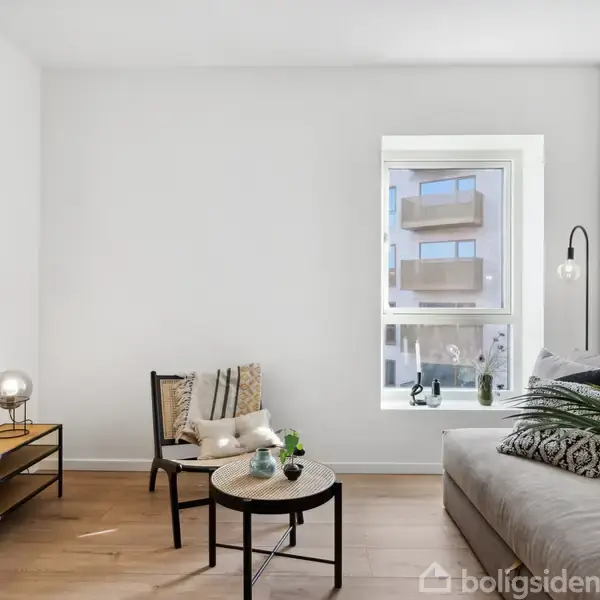 En lys stue har en sofa med puder til højre, en stol og et lille bord i midten. I baggrunden ses en reol med planter, en lampe og plakat.