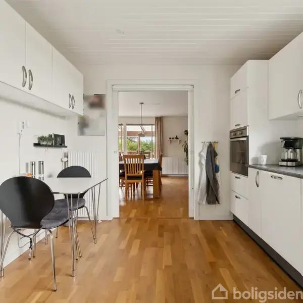 Køkken med hvidt interiør og trægulv, hvor et lille bord med to stole står op ad væggen. I baggrunden ses åbningen til en spisestue med et spisebord og stole.