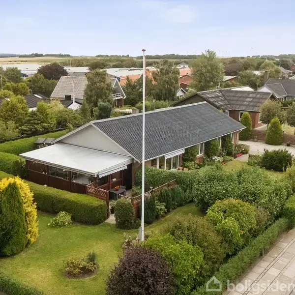 En enfamiliehusejendom med mørkt tag er omgivet af velplejede hække og grønne buske i et villakvarter. En flagstang står foran huset, som ligger ved en vej.