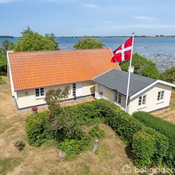 Hus med rødt tegltag og dansk flag vajende fra flagstang. Beliggende ved kysten med havudsigt og omgivet af grønt haveområde samt spredte buske og træer.