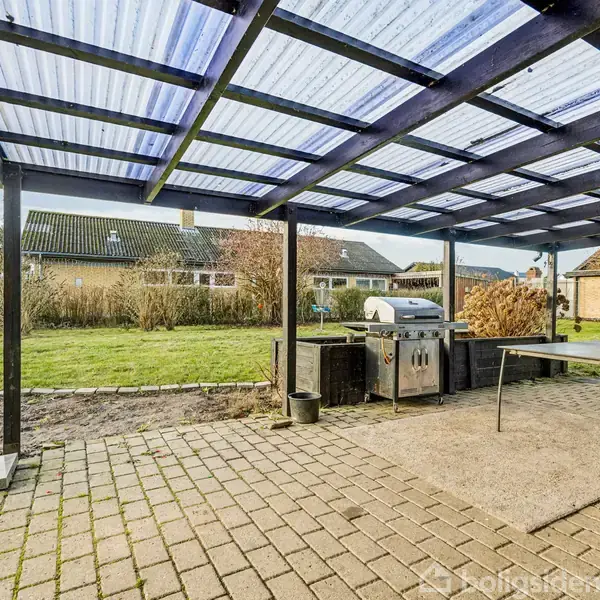 Grill står på en overdækket terrasse med flisebelægning og bord. Omkring er grønne plæner og bungalowlignende huse. Parasollen er samlet, mens en rolig atmosfære præger haven.