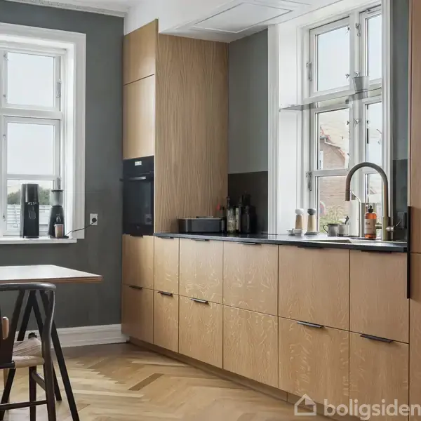 Et moderne køkken med lyse træskabe langs en væg. Der er en sort bordplade med forskellige køkkenredskaber på. Et stort vindue giver naturligt lys.