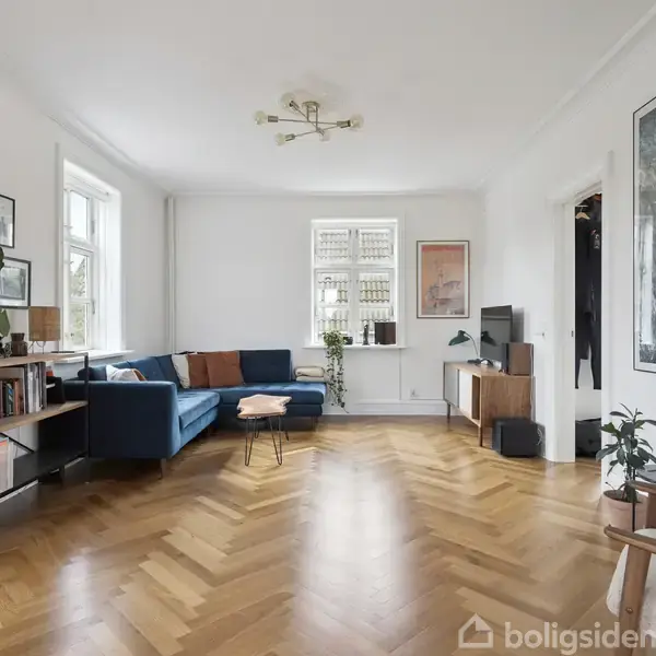 En stue med en blå sofa i hjørnet, en reol med bøger til venstre og et TV på et skab til højre. Trægulv og kunst på væggene.