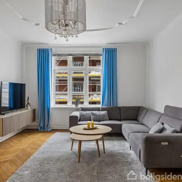 En grå sofa står i stuen ved et rundt sofabord med lys. En fladskærm hænger på en væg, lysindfald fra vindue med blå gardiner. Rummets gulv er trægulv.