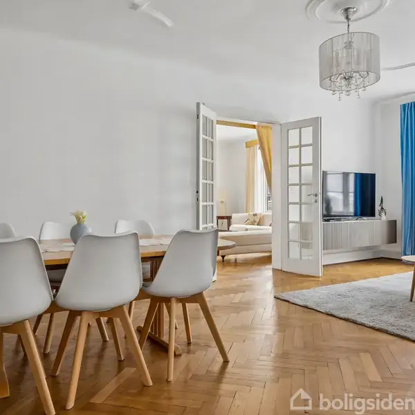 Spisebord med seks hvide stole står på trægulv. I baggrunden er der en stue med sofa og tv. Væggene er hvide med blå gardiner ved vinduet. Skilt viser “Værkult!” og “Tacofredag".