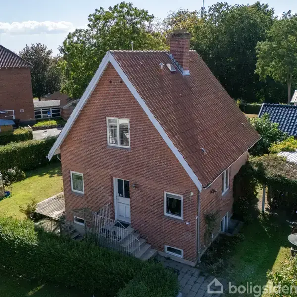 En rød murstensvilla med en stejl tagryg står omgivet af en velplejet have med hæk. En lille terrasse med bord og stole ses ved siden af huset.