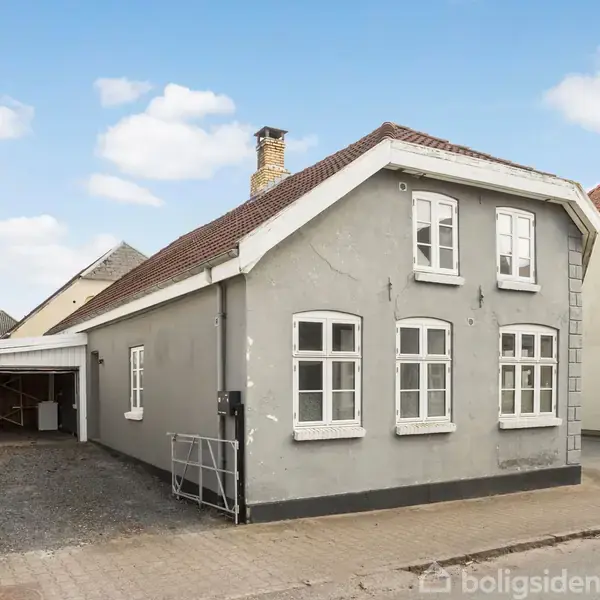 Et ældre, gråt hus står stille på en brolagt gade, omgivet af andre huse. Det har hvide vinduer og en brun teglstenstag. Der er en parkeringsplads ved siden af.
