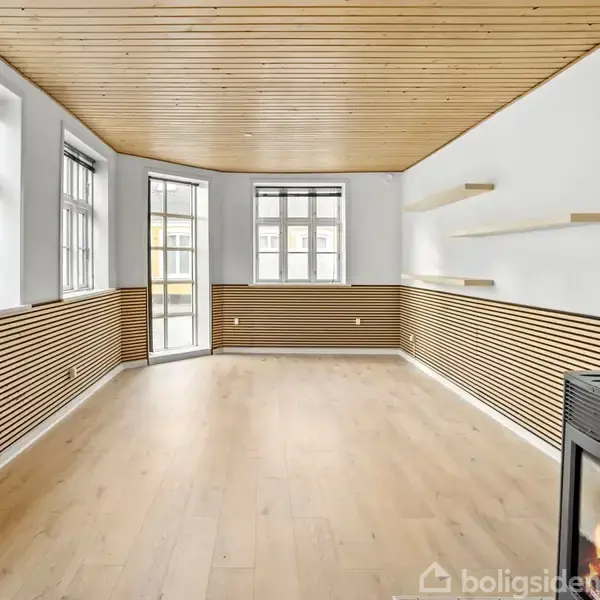 Stue med trægulv og træloft, vægge dekoreret med vandrette trælameller, har indbyggede hylder. Brændeovn i højre hjørne. Store vinduer og glasdør giver lysindfald.