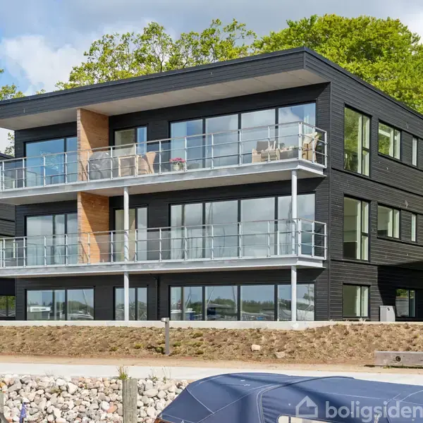 En moderne sort bygning med store glasfacader står ved en vandkant. Bygningen har to altaner, og omgivelserne inkluderer grønne træer og en stenmuret kaj.