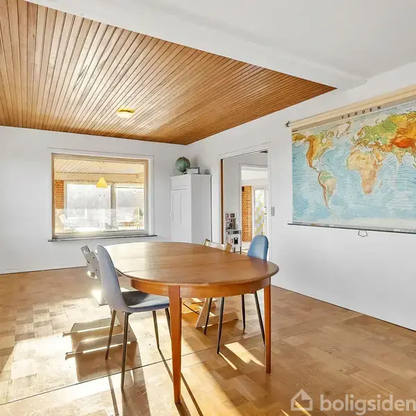 Rundt bord med tre stole står i et rum med trægulv og træloft. En verdenskortplakat hænger på væggen. Rummet har store vinduer og hvide vægge, med en afslappet atmosfære.