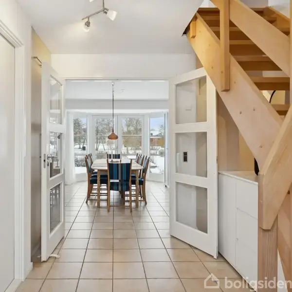 En gang med beige flisegulv fører til en spisestue med et bord og seks stole, omgivet af store vinduer. En trætrappe går op til højre. Ingen tekst fundet.