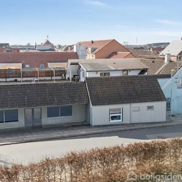 Et langt hus med mørkt tag ligger ved en stille vej i en landsbyomgivelser. Der er flere vinduer og en dør på facaden, omgivet af andre bygninger.
