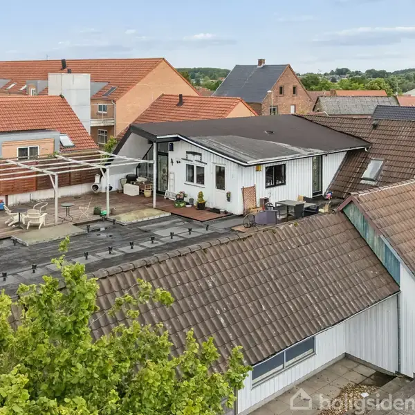 En bygning med fladt tag og hvid facade, omgivet af andre hustage. Der er en terrasse med havemøbler og planter. Omkringliggende røde tage og grønne træer kan ses i baggrunden.