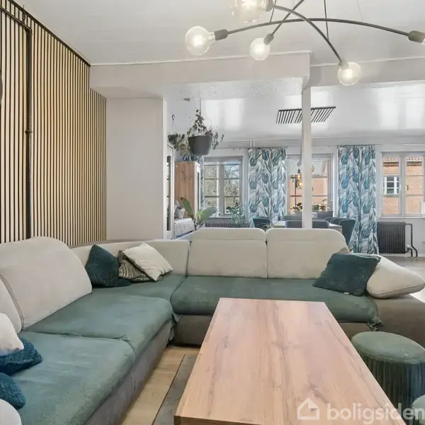 Stor sofa står i centrum, flankeret af træbord, under moderne loftlampe. Bagved ses et vinduesparti med gardiner. Væg bliver udsmykket med stor ur og lodrette paneler.