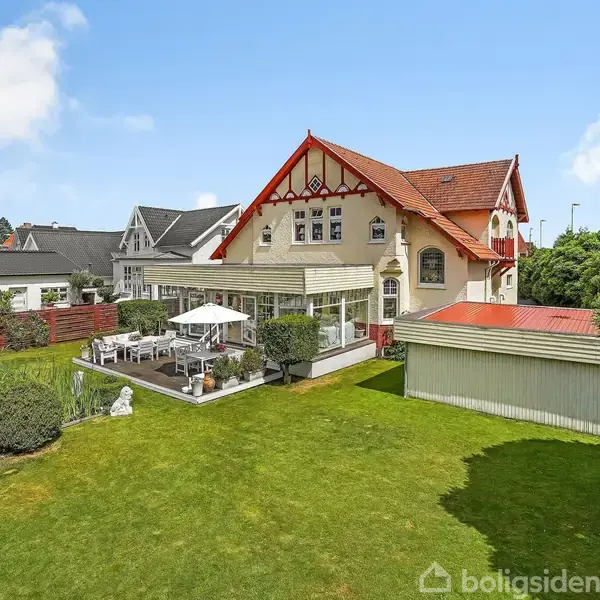 Et charmerende hus med stort, rødt tegltag står i en velplejet have. Terrassen ved siden af er udstyret med hvide møbler og parasoller, omgivet af buske og græsplæne.