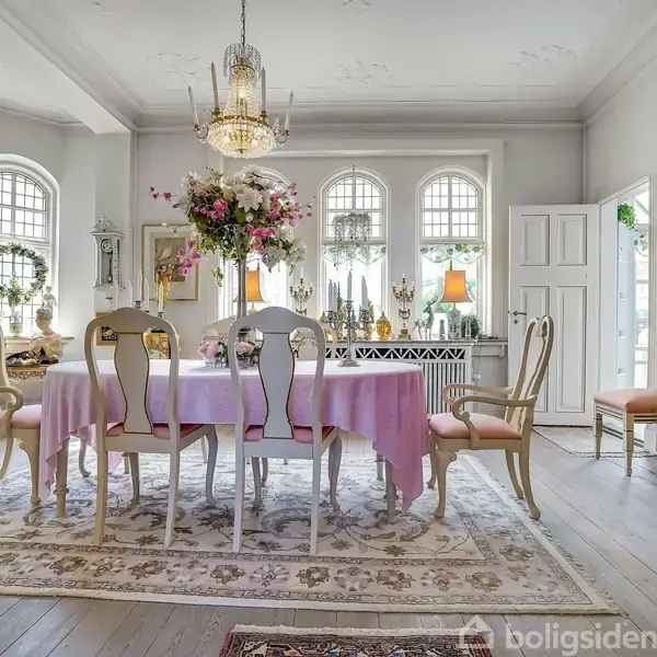 Spisestue med stort bord dækket af lyserødt dug og blomsterdekoration. Omgivet af elegante stole, lysekrone og vinduer med buede rammer. Lamper og pynt pryder rummet.