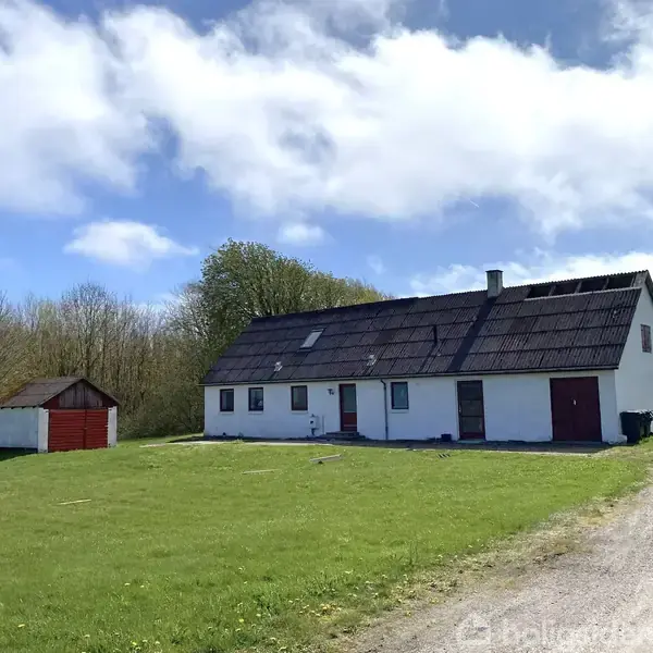 Hvidt hus med rødt tag på stor græsplæne, ved siden af et rødt skur. Omkranset af træer under blå himmel med hvide skyer.