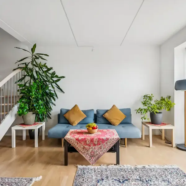 Sofa med to puder står foran et trappegelænder; to planter på sidder på små borde. Et stort vindue med udsigt; et tæppe på gulvet.