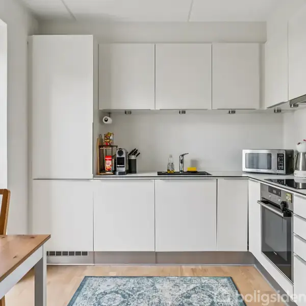 Et køkken med hvide skabe og apparater. En kaffemaskine, elkedel og mikroovn er på bordpladen. Et spisebord med en plante står ved vinduet. En lys tæppe ligger på gulvet.