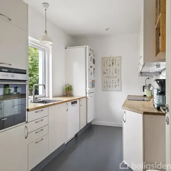 Et lyst køkken med hvide skabe, træbordplader og moderne apparater. Køkkenet har en vask ved et vindue med udsigt til grønne træer. En plakat hænger på muren.