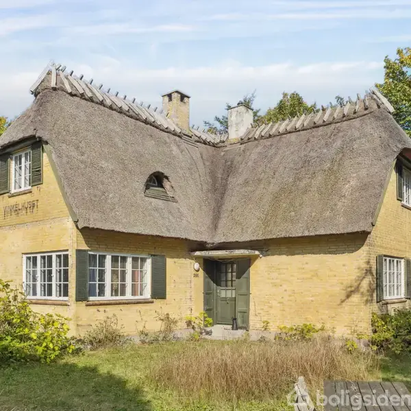 Stråtækt hus med gule mursten står stille i en frodig have omgivet af træer. Datoen "1928-1957" er skrevet på facaden.