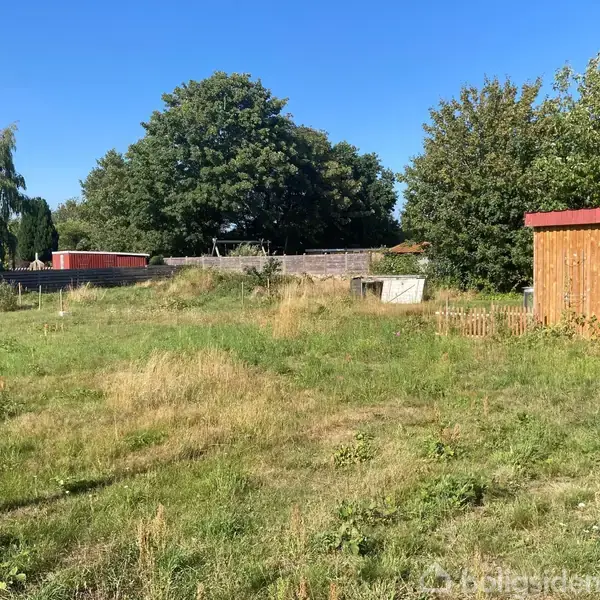 Et lille træskur med en rød tagkant står på en græsplæne. Der er også nogle betonblokke ved siden af skuret. Baggrunden består af buske og træer.